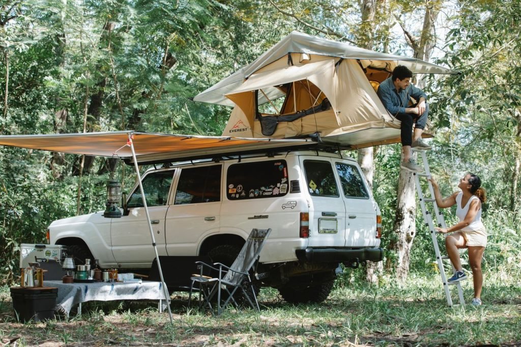 Full body of young male camper relaxing in tent placed on SUV car roof and chatting with Hispanic girlfriend climbing ladder during trip in nature on sunny day